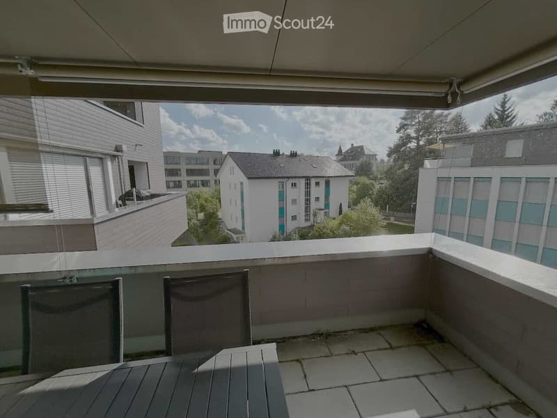 Balcony with wooden decking, glass railing, and a view of surrounding buildings and greenery.