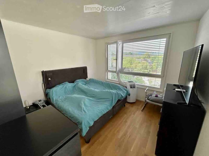 A well-lit bedroom with wooden flooring, a large window with blinds, a bed with a blue duvet, a nightstand, a television, an air conditioner, and a small chair.