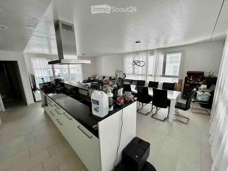 Modern kitchen with white cabinets, black countertops, and a central island. There is a dining area with a white table and black chairs, and various kitchen appliances and items are visible on the counters.
