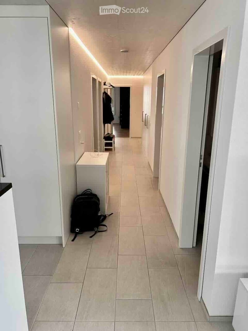 Clean, modern hallway with white walls, tiled floor, and built-in storage units. A backpack is placed on the floor near the left wall.