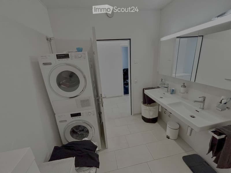 Modern bathroom with a double sink, large mirror, and a stacked washer/dryer unit. The space features white tiles, a small trash bin, and a towel on the floor.