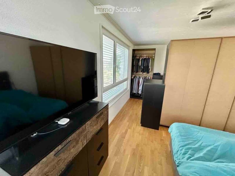 Modern bedroom with a large flat-screen TV on a wooden dresser, a neatly made bed with blue bedding, and a built-in wardrobe with hanging clothes.
