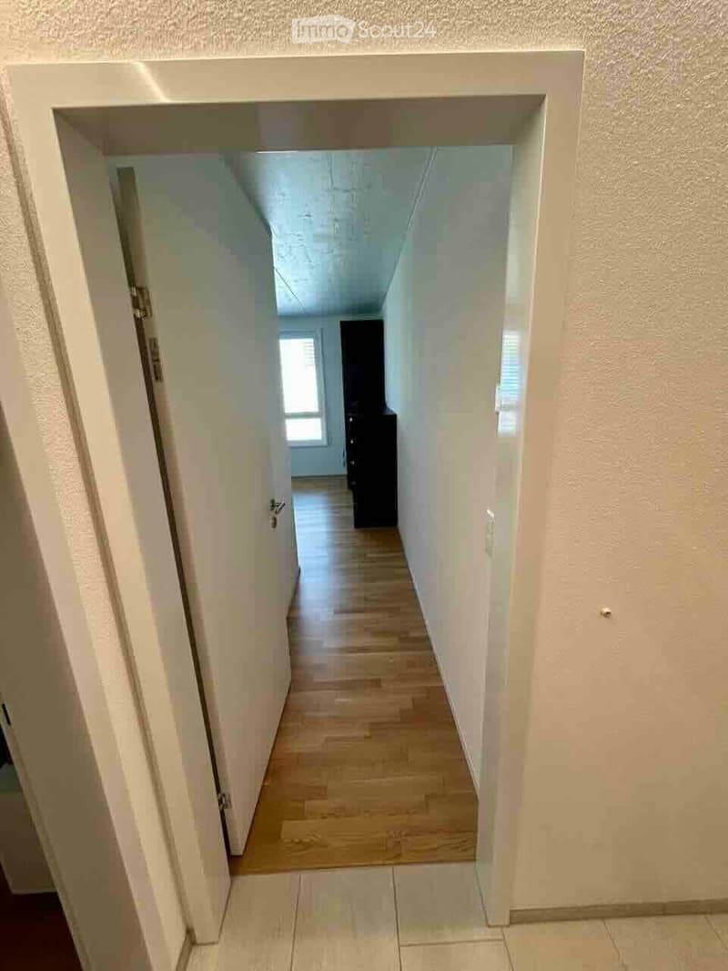 A clean, well-lit hallway with wooden flooring and white walls, leading to a room with a visible piece of furniture.