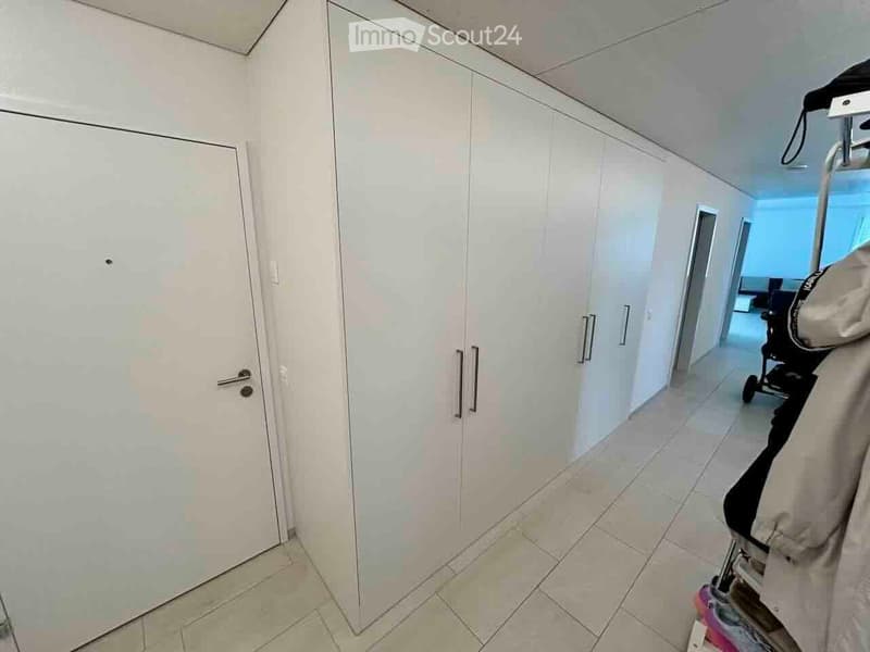A clean, modern hallway with white walls, tiled flooring, and built-in white closet units with silver handles. A stroller is visible in the background.