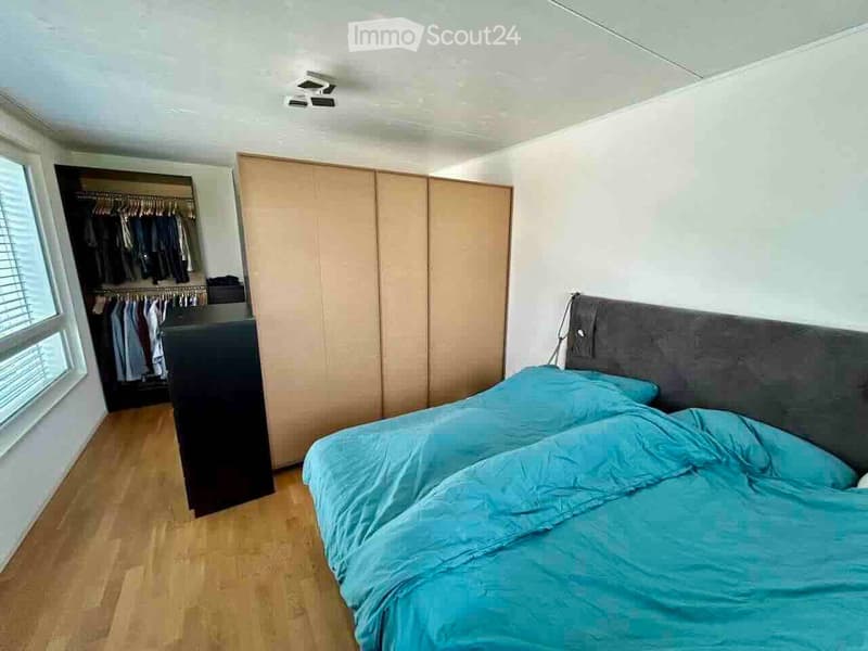 Modern bedroom with a large bed covered in blue bedding, a dark headboard, a built-in wardrobe, and a closet with hanging clothes. The room features wooden flooring and large windows with blinds.