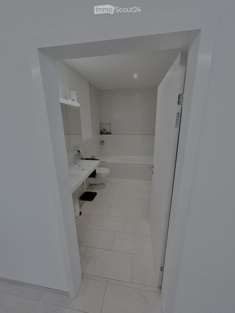 Modern bathroom with white tiled walls and floor, featuring a sink, toilet, bathtub, and a mirrored cabinet.