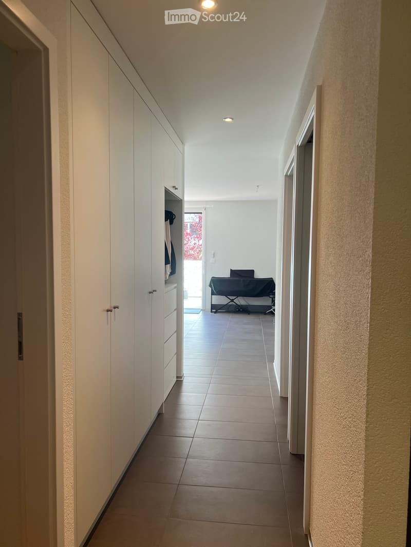 A modern hallway with tiled flooring, white walls, built-in white cabinets on one side, and a small room visible at the end containing a massage table.