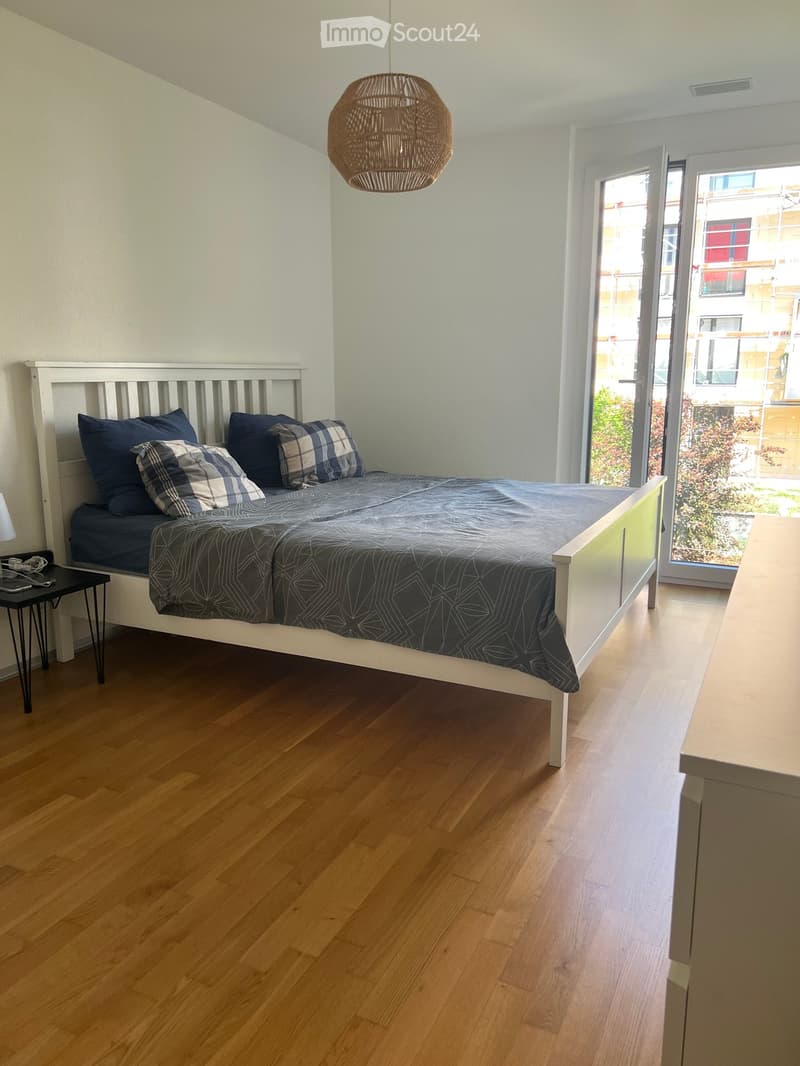 Modern bedroom with wooden flooring, white walls, a single bed with dark bedding, a nightstand with a lamp, and a wicker pendant light hanging from the ceiling. A large window with a view of the city is visible.