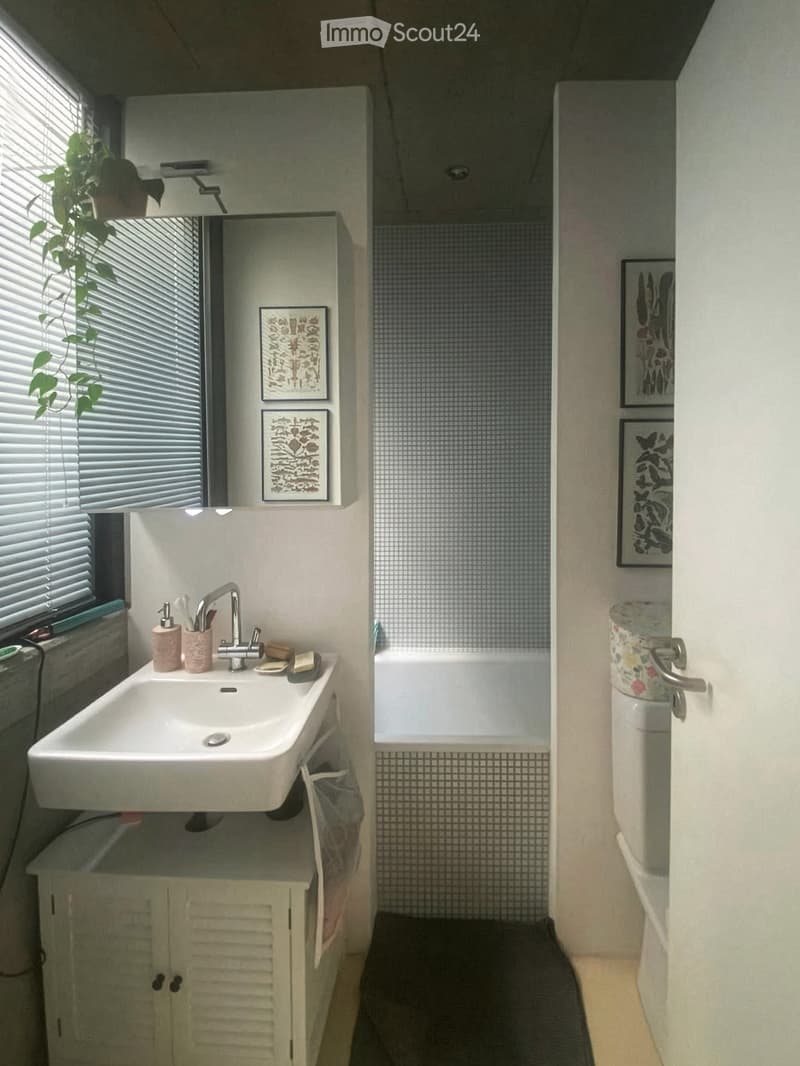 Bathroom with a white sink, faucet, and cabinet, a bathtub with tiled walls, a toilet, and framed artwork on the walls. A window with blinds and a potted plant are visible.