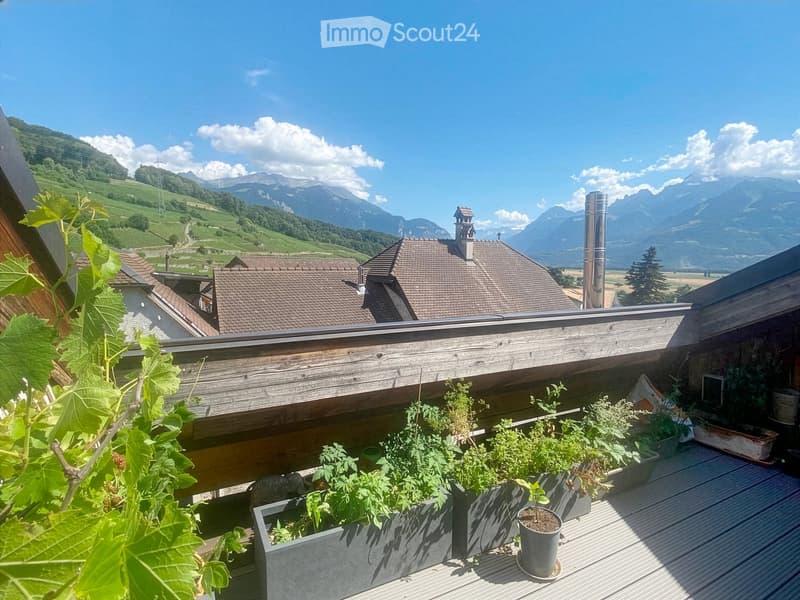 A wooden deck with potted plants and vines, overlooking a scenic valley with mountains and houses in the background.