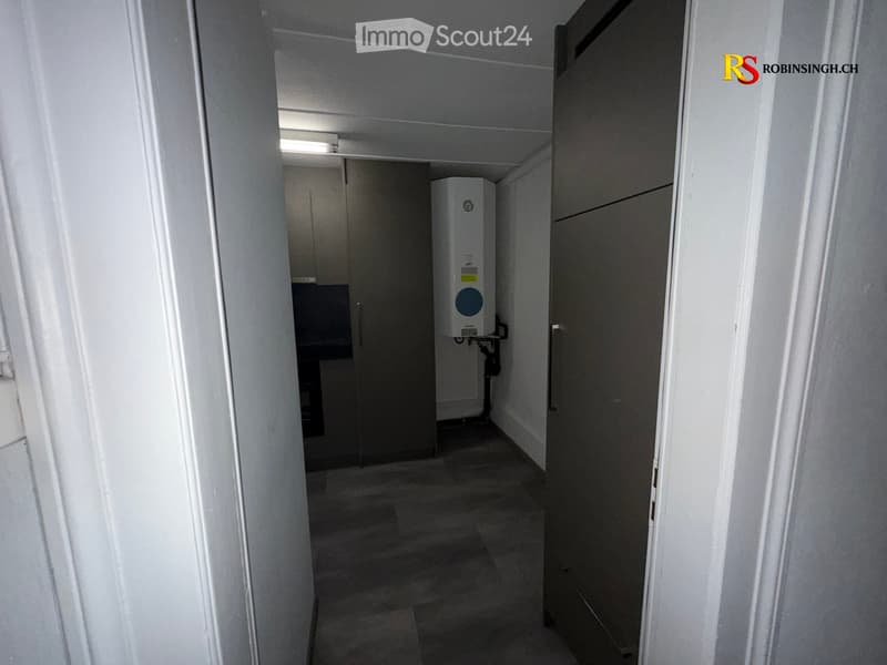 A narrow, dimly lit hallway with a tiled floor, white walls, and a water heater unit visible in the background.