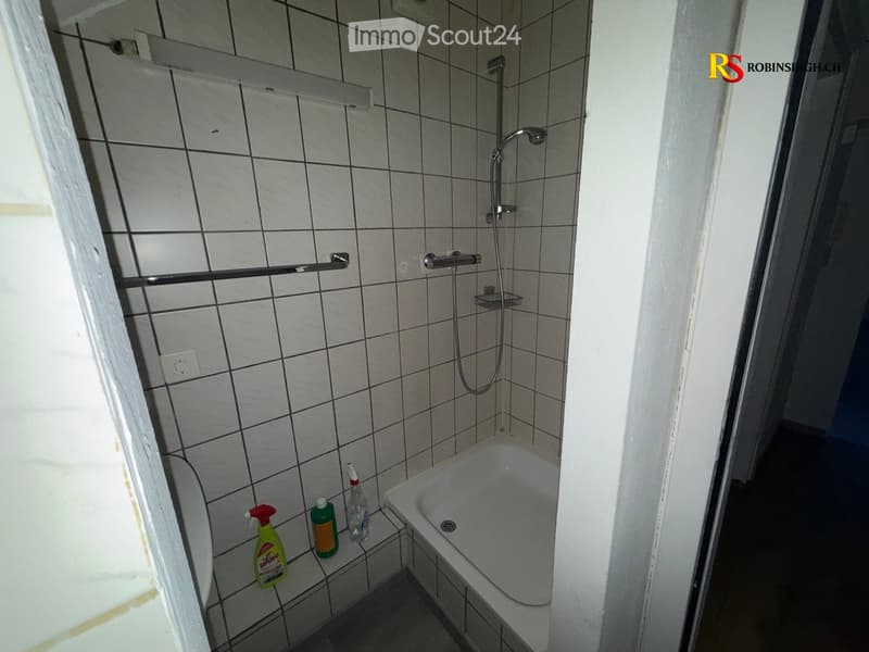 A small bathroom with a bathtub, toilet, and shower. Cleaning supplies are placed on the edge of the bathtub. The walls are tiled with white tiles and black grout.