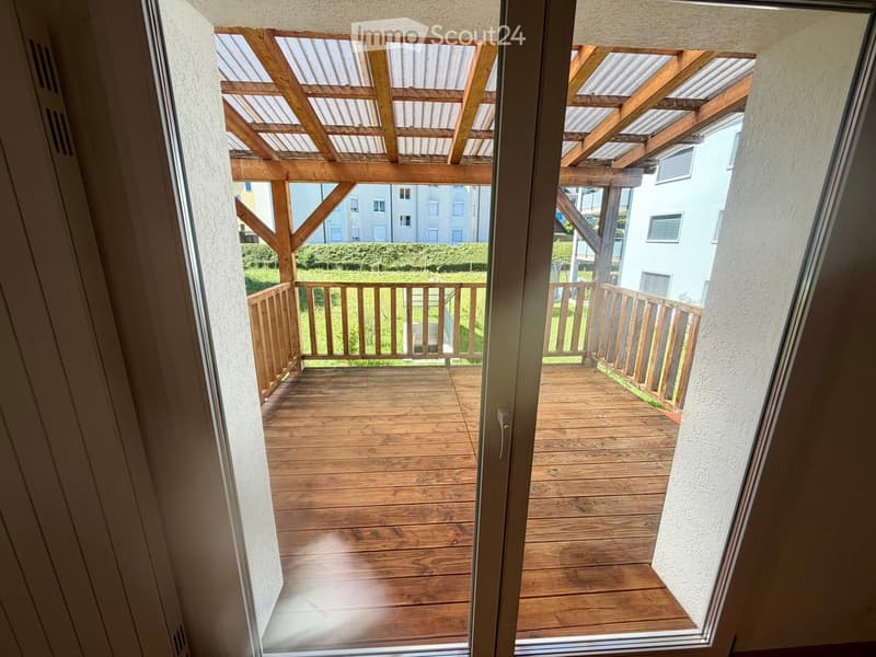 Wooden balcony with a slatted wooden roof and wooden railing, overlooking a green lawn and neighboring buildings.