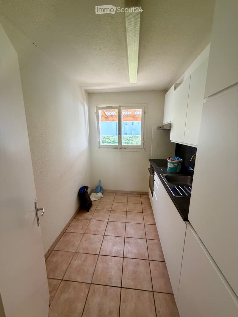 Empty kitchen with tiled floor, white cabinets, sink, and oven. A small window with blinds allows natural light.