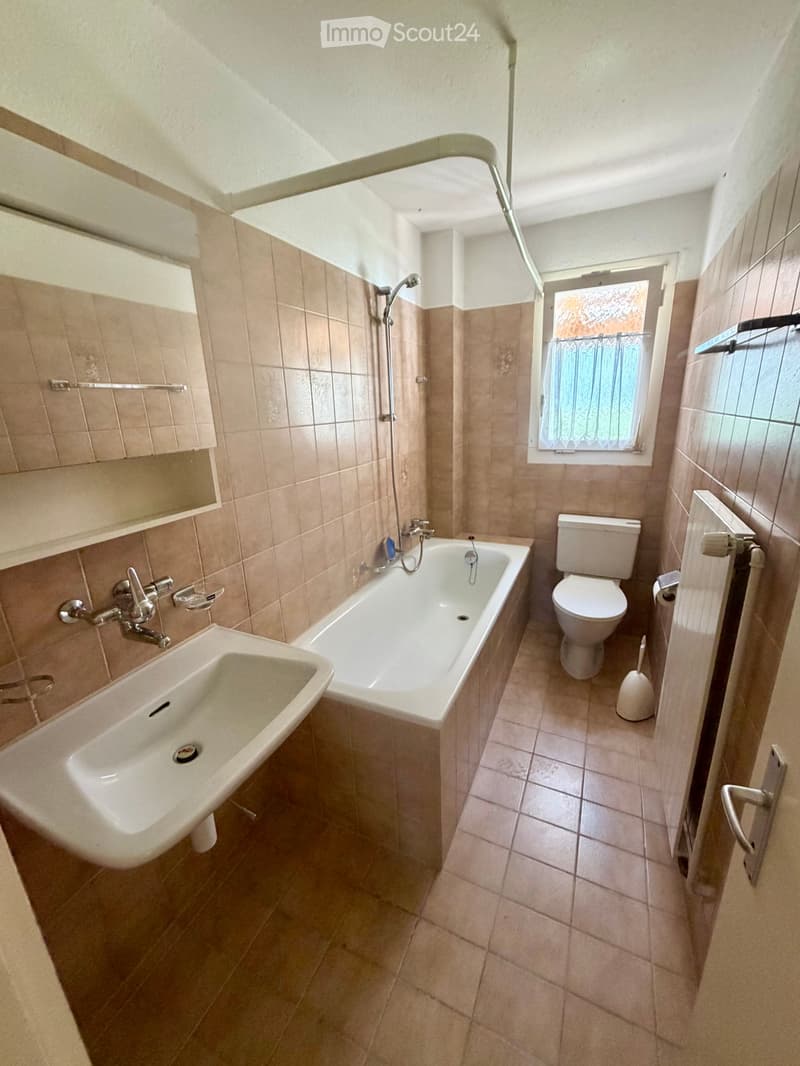 Bathroom with beige tiled walls and floor, featuring a sink, bathtub, toilet, and showerhead. A window with a decorative glass panel is visible.