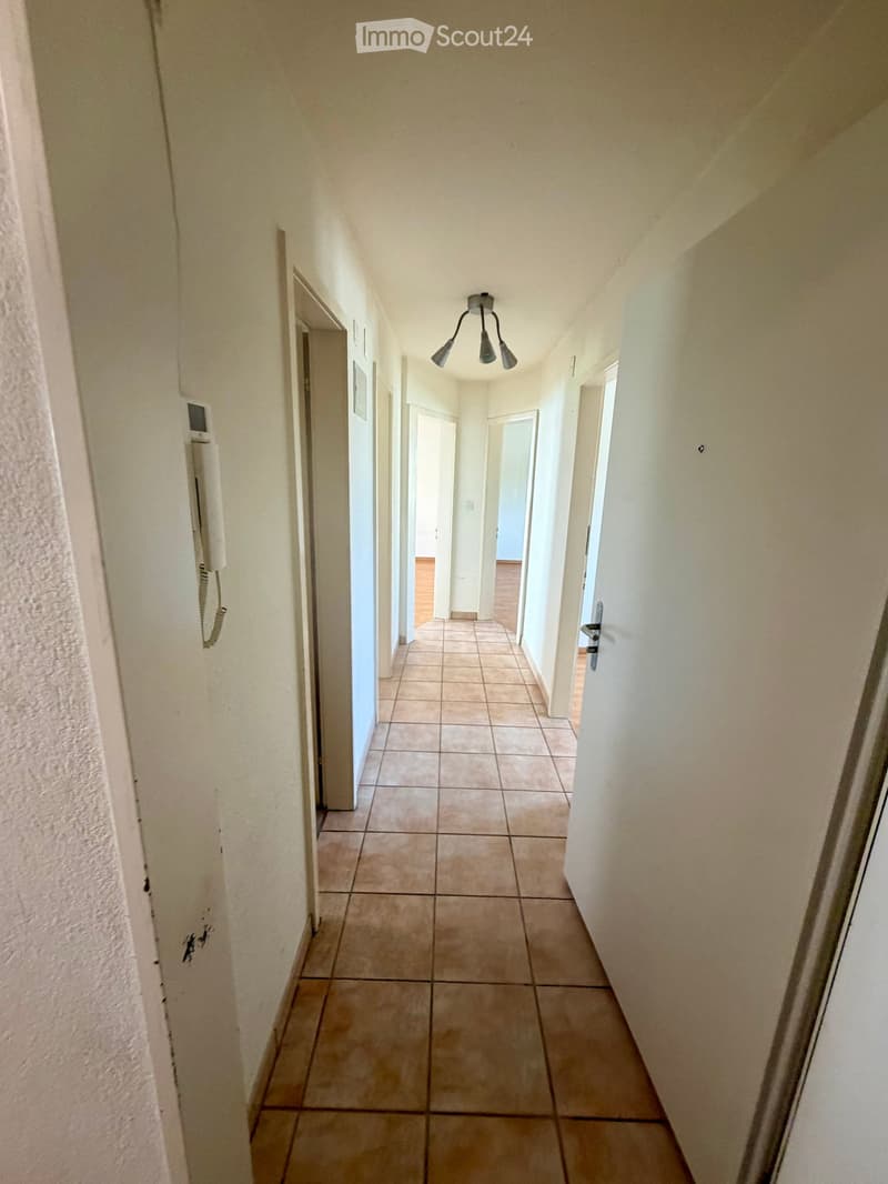 A narrow hallway with tiled flooring, white walls, and several doors leading to other rooms. A light fixture hangs from the ceiling, and a phone is mounted on the left wall.