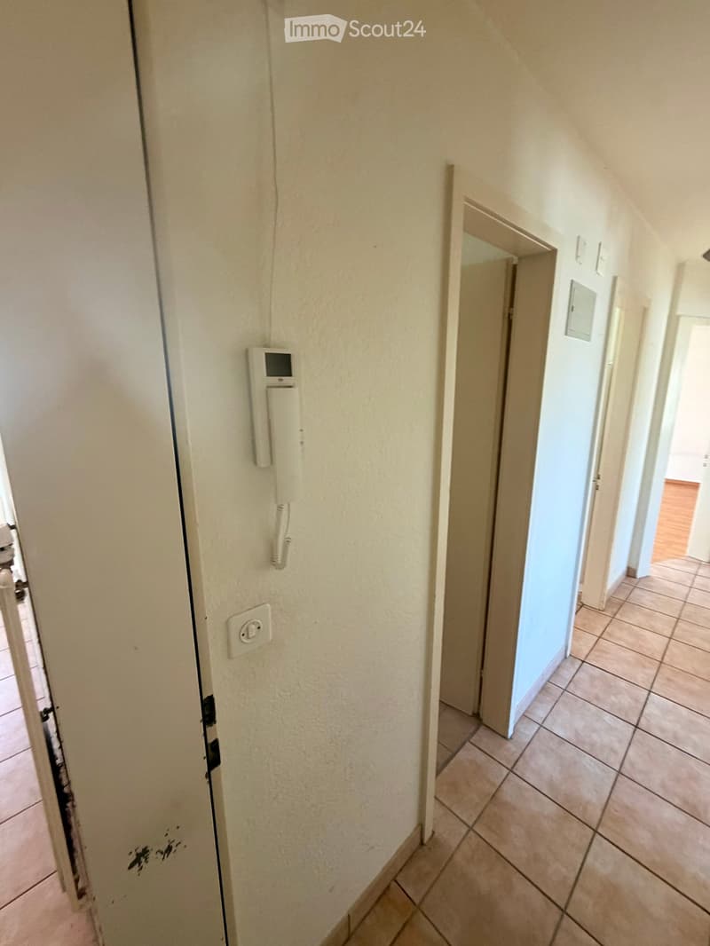 A narrow hallway with beige tiled flooring, white walls, and a door on the left. A wall-mounted intercom system is visible on the left wall, and an open doorway leads to another room with wooden flooring.
