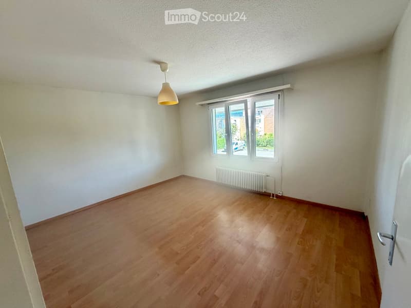 Empty room with wooden flooring, white walls, a window with blinds, a radiator, and a hanging light fixture.