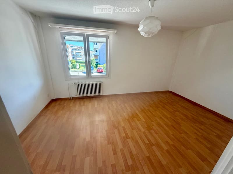 Empty room with wooden flooring, white walls, a radiator, and a large window offering a view of the street and buildings outside.