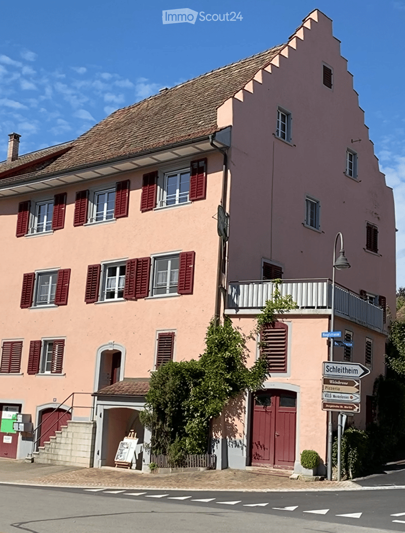 A two-story pink building with red shutters and a tiled roof, featuring a small balcony on the upper floor and a signpost nearby.