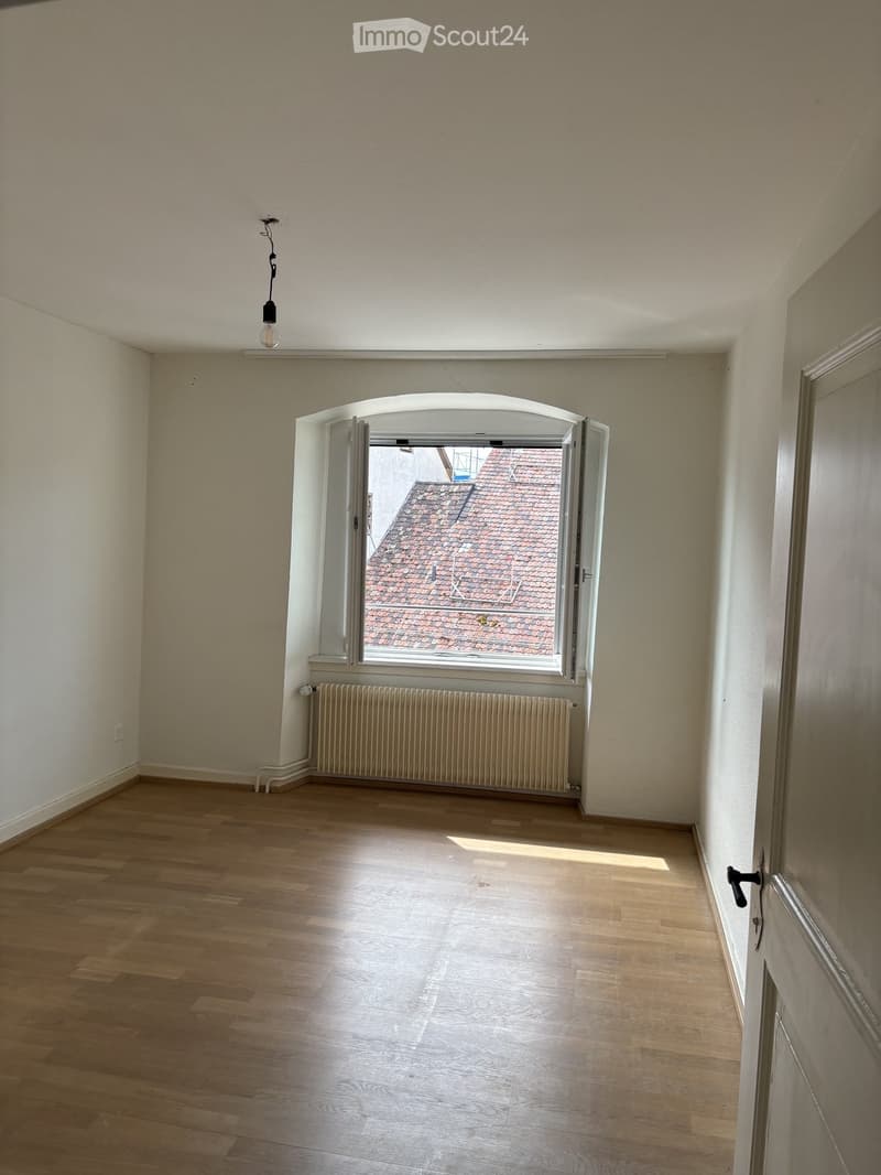 Empty room with wooden flooring, white walls, a radiator, and a large arched window offering a view of a tiled roof.