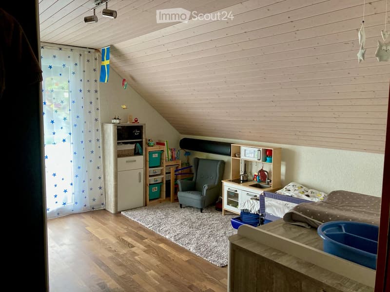 A small room with wooden floor, white walls, a bed, a chair, a desk, a cabinet, a rug, and a window with a curtain.