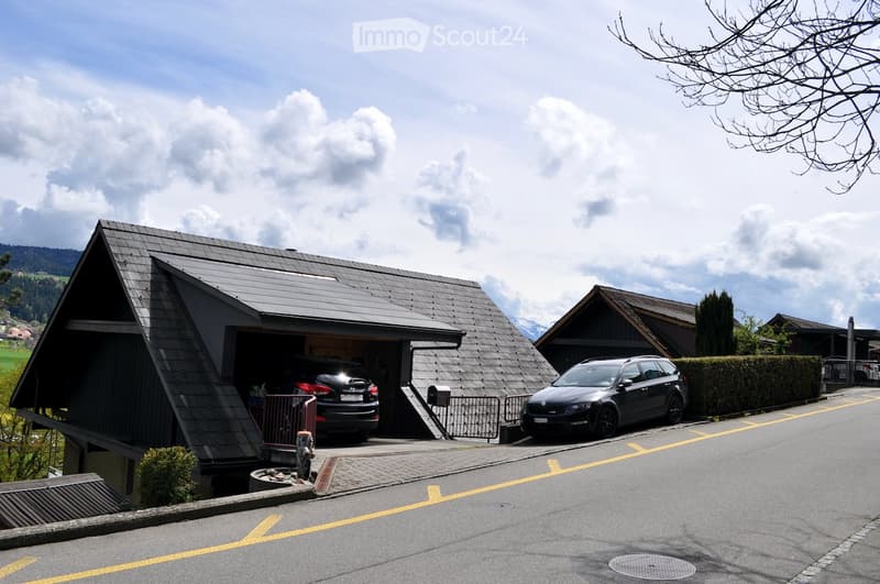 House with a garage, two cars parked in the garage, another car parked on the street, surrounded by other houses and greenery.