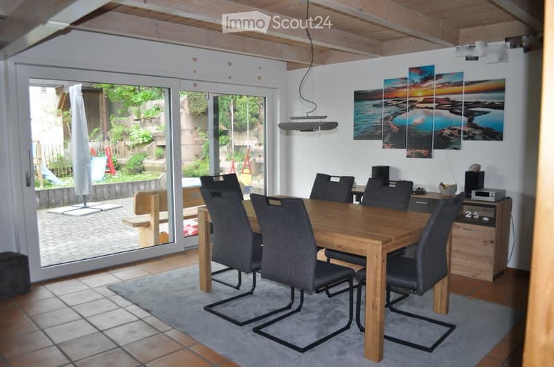 Dining room with wooden table, gray chairs, sliding glass door, wooden bench, tiled floor, carpet, wall with paintings, wooden cabinet, speakers, and a ceiling lamp.