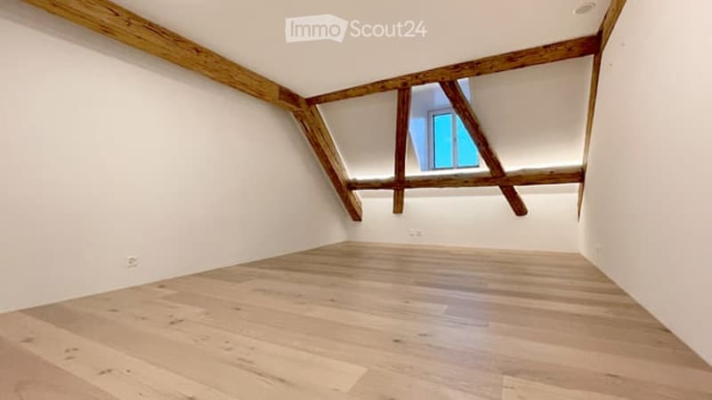 Empty attic room with wooden beams and exposed wooden floor, featuring a skylight window.