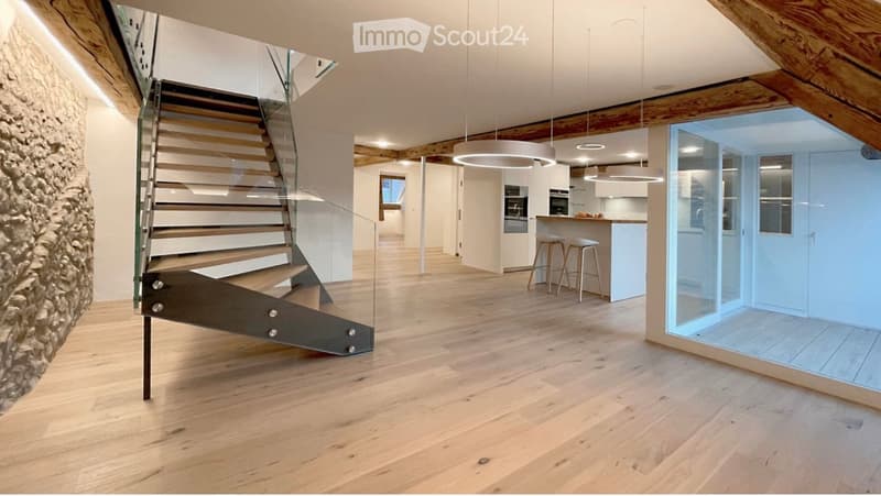 Modern interior with wooden flooring, a sleek staircase with glass railing, pendant lights, and a kitchen area with a small island and stools.