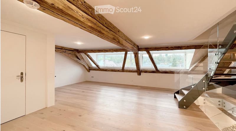 Bright, open attic space with exposed wooden beams, skylights, and a modern glass staircase leading to another level.