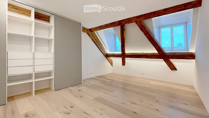 Empty attic room with exposed wooden beams, skylight windows, white walls, and light wooden flooring. Features built-in white shelving and a closet.