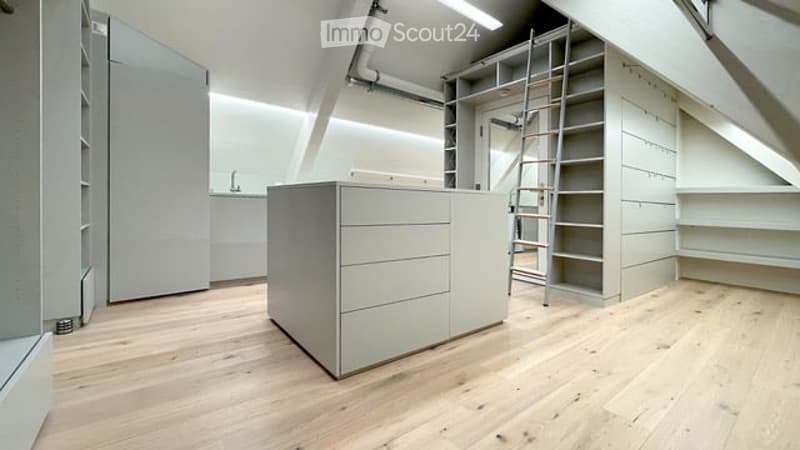 Bright, minimalist attic space with exposed wooden beams and slanted ceilings. Features built-in white cabinetry, a central island with drawers, and a ladder leading to an upper level.