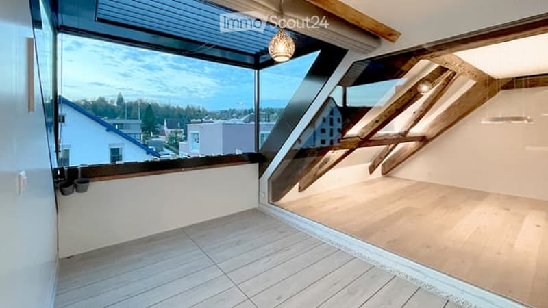 Bright, open loft-style room with large windows offering a scenic view of the city and surrounding landscape. The space features wooden flooring and exposed wooden beams, creating a modern and airy atmosphere.