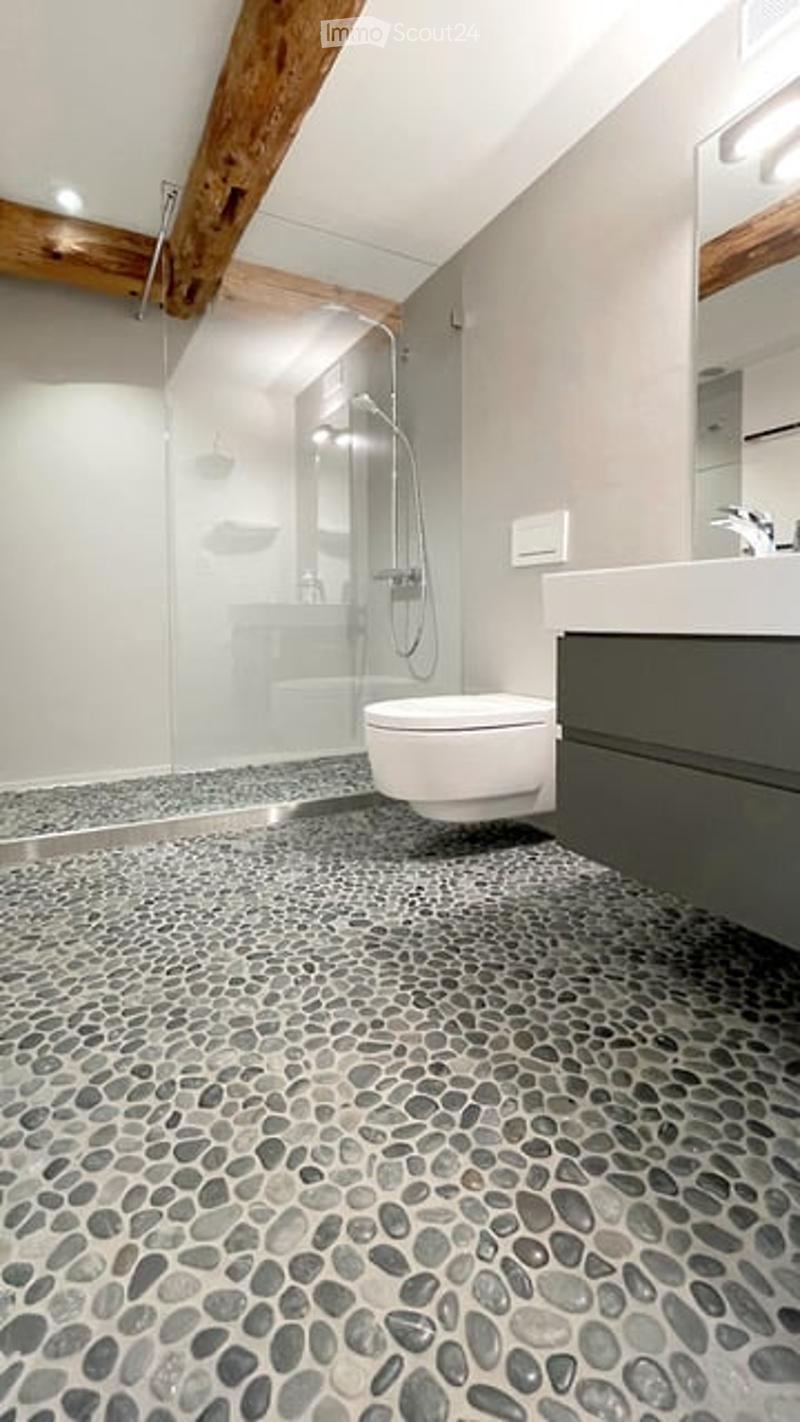 Modern bathroom with a pebble tile floor, white toilet, sink with dark cabinet, glass-enclosed shower, and exposed wooden beams on the ceiling.
