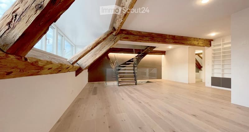 Bright, open loft space with exposed wooden beams, light wooden flooring, and a modern staircase leading to an upper level. Large windows allow natural light to fill the room.