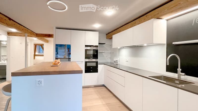 Modern kitchen with white cabinets, a central island, stainless steel sink, and built-in ovens. Exposed wooden beams on the ceiling and recessed lighting.