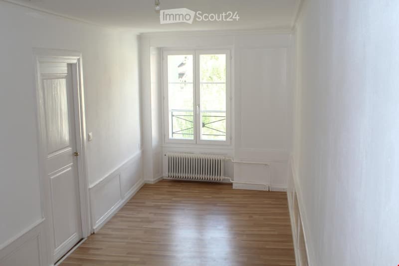Empty room with white walls, wooden floor, a window with a radiator below, and a door on the left side.