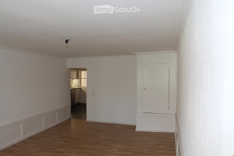 Empty room with wooden flooring, white walls, and a single light fixture. A doorway leads to a small kitchen area with cabinets and a sink.