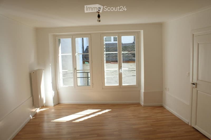 Empty room with wooden flooring, white walls, large windows, and a radiator on the left side.