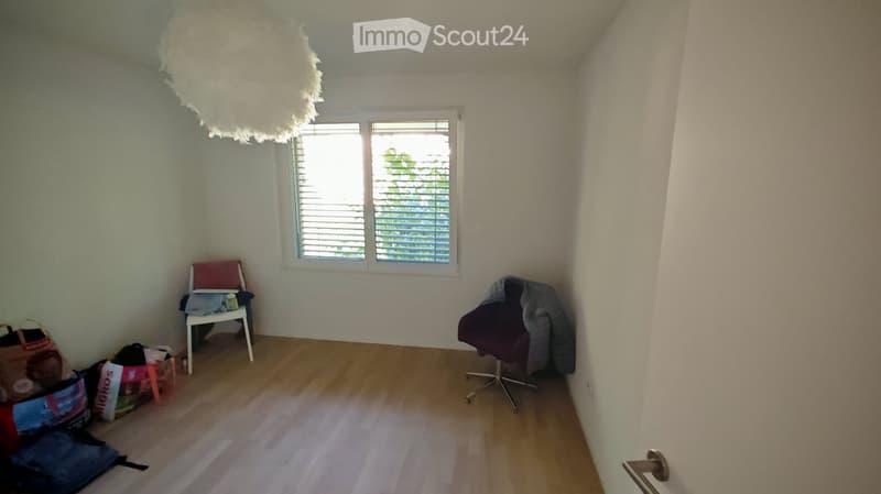 Empty room with wooden flooring, white walls, and a large window with blinds. A single chair and a small stool are present, along with a decorative lamp hanging from the ceiling.
