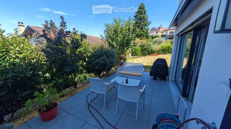 Balcony with tiled floor, white wicker furniture including a sofa, table, and chairs, a covered grill, and a potted plant.