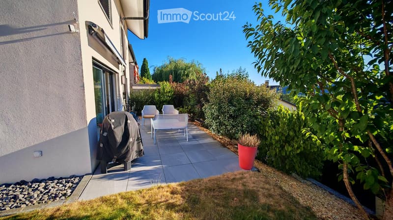 A modern outdoor patio area with a tiled floor, white chairs, a small table, and a red flower pot, surrounded by greenery and a house with large windows.