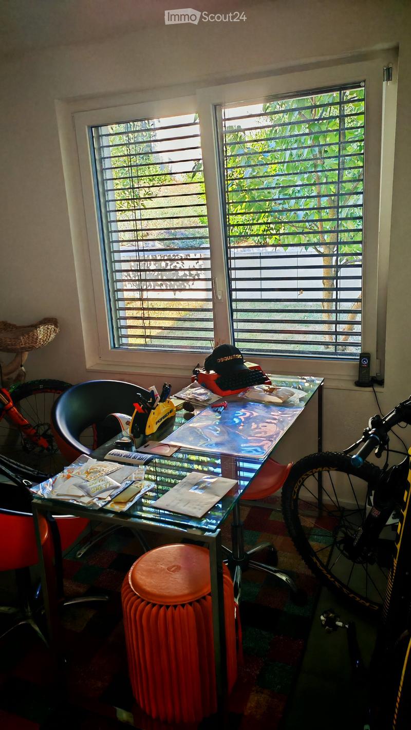 A small, furnished room with a glass table, a black office chair, a red plastic stool, and a bicycle leaning against the wall. The room has two windows with metal bars and a colorful carpet.