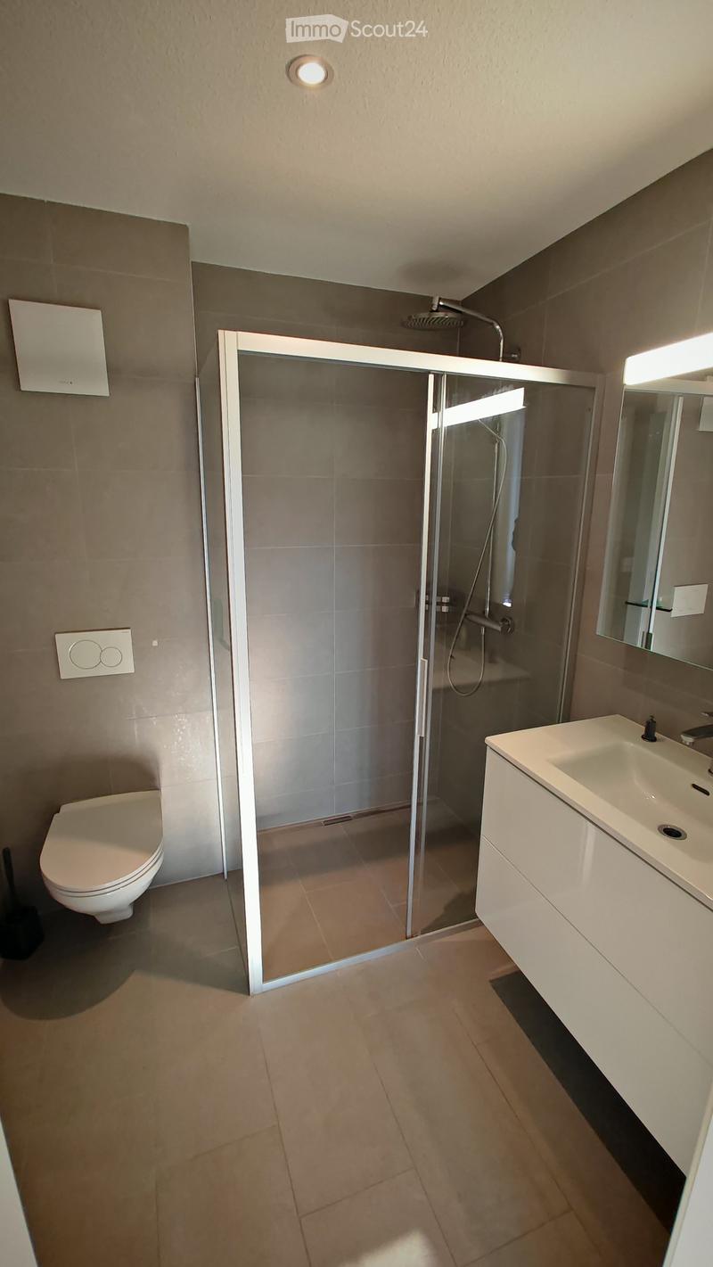 Modern bathroom with a glass-enclosed shower, toilet, sink, and mirror. Light-colored tiles and a sleek design.