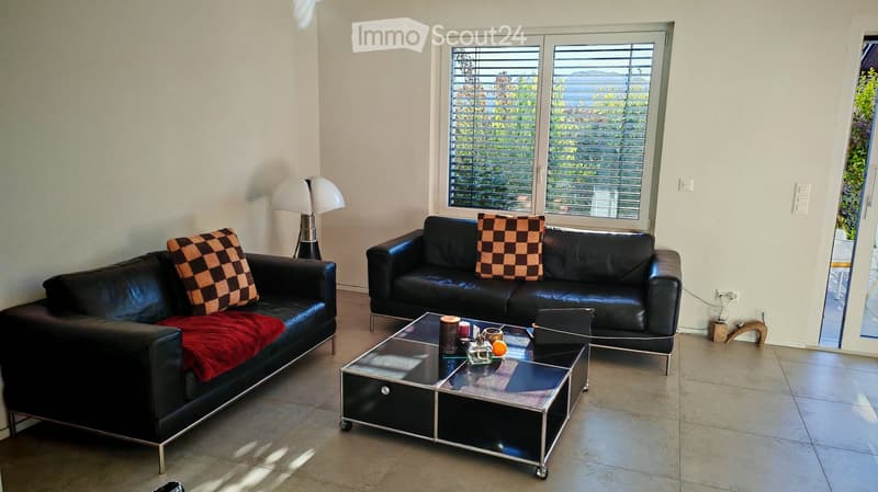 Modern living room with two black leather sofas, a glass coffee table, and a floor lamp. The room features large windows with blinds and a view of greenery outside.