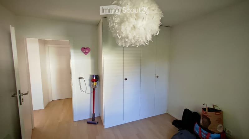 Empty room with light green walls, wooden flooring, white closet, and a decorative heart-shaped wall hanging. A vacuum cleaner is placed near the closet, and a shopping bag is on the floor.