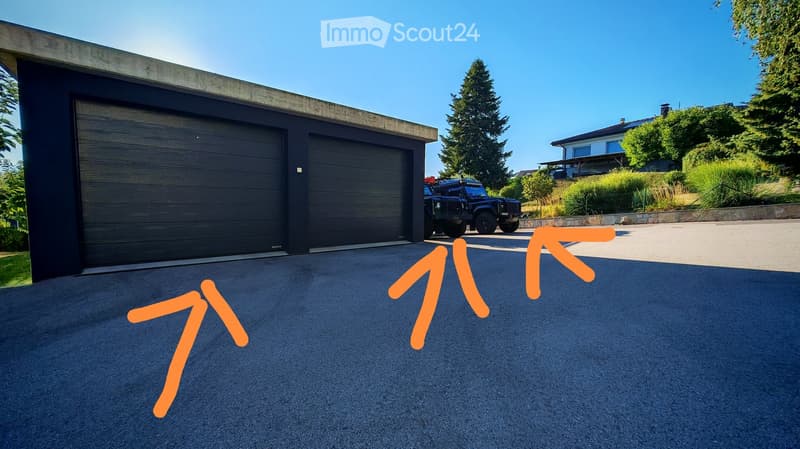 Modern two-car garage with dark wooden doors, situated on a paved driveway with a black SUV parked nearby. The surrounding area includes greenery and a neighboring house visible in the background.