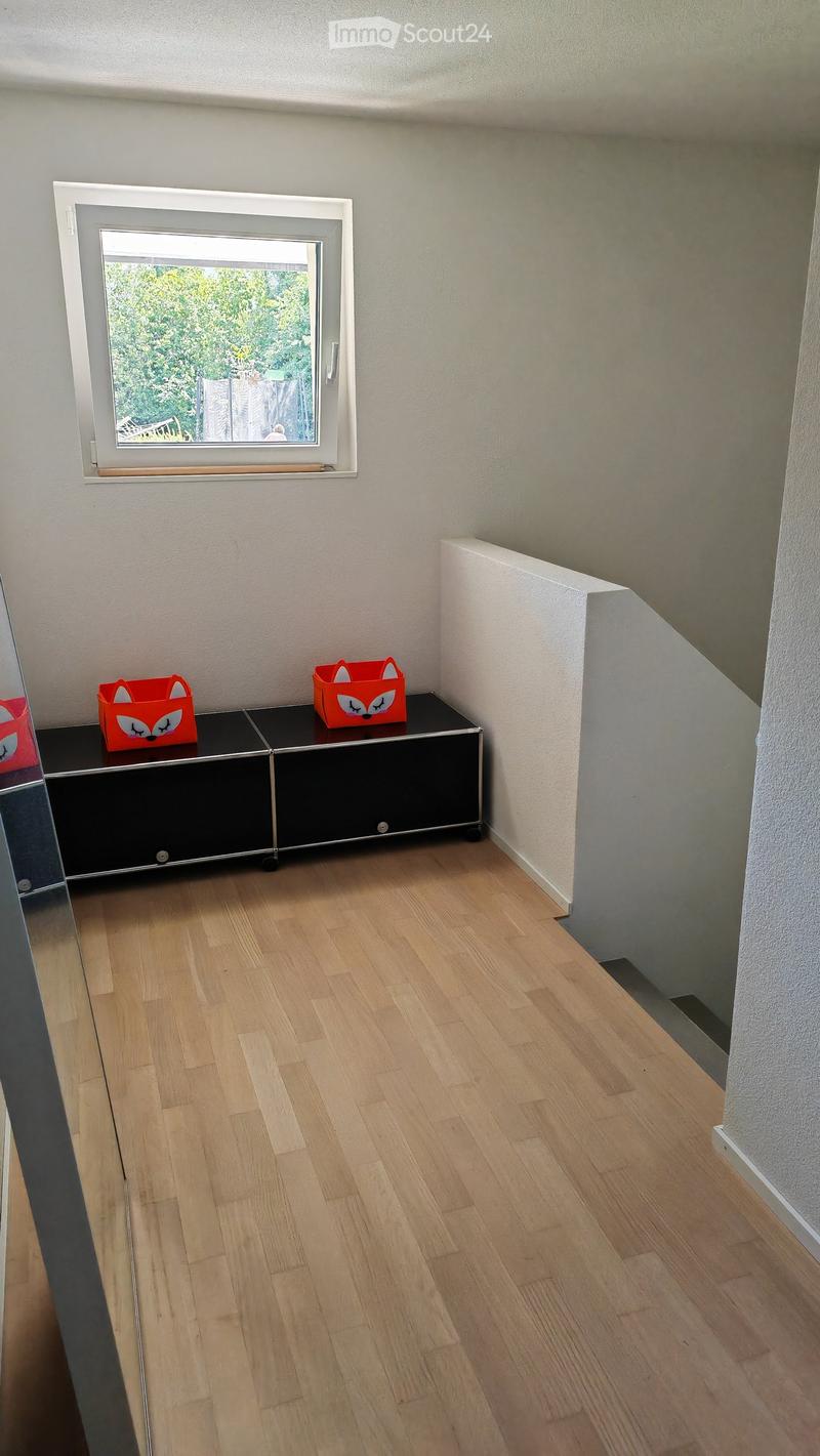 A small, unfurnished room with light wooden flooring, white walls, and a window showing greenery outside. A black cabinet with red fox-shaped toys on top is visible.
