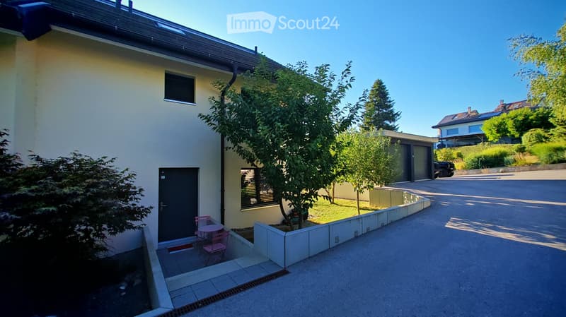 A modern house with a small garden area featuring a table and chairs, surrounded by trees and a paved driveway.